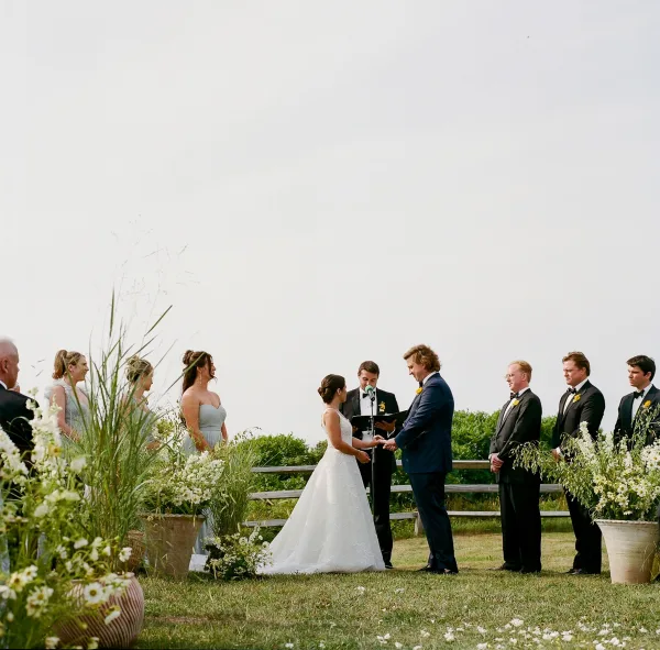 Wedding vows as bride and groom hold hands while officiant reads into a microphone before bridesmaids and groomsmen on a lawn
