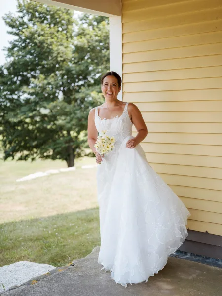 Bridal portrait of a smiling bride holding a daisy bouquet, showing her wedding ring in a white floral applique dress on a yellow porch.
