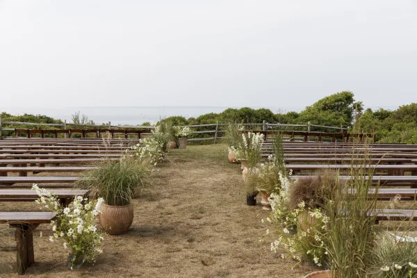 Ceremony setup with outdoor ceremony seating on wood benches, lined with white aisle florals and terracotta pots overlooking the ocean view