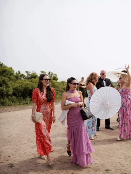 Wedding guests in wedding guest outfits walk along an outdoor dirt path, wearing colorful dresses and tuxedo amid greenery under open sky