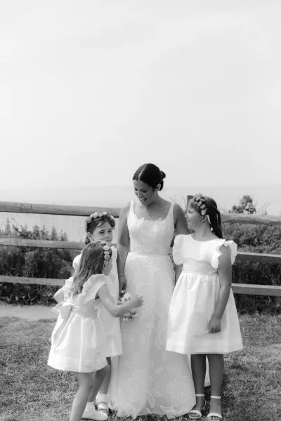Bride with flower girls in white flower girl dresses, laughing on a grassy lawn by a wood fence with ocean view, holding a bouquet