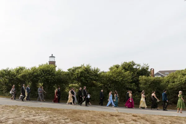 Wedding guests in wedding guest attire walk along a roadside toward a lighthouse, wearing suits, ties, sunglasses, and gowns under overcast sky