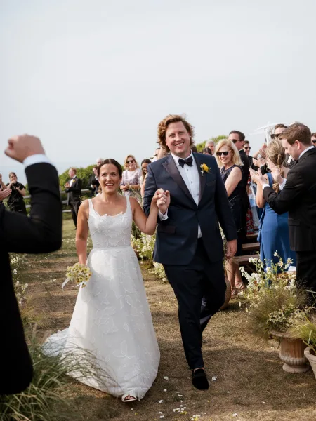 Wedding recessional as bride and groom walk the aisle holding hands, bouquet raised, guests cheering on a coastal lawn with ocean horizon behind