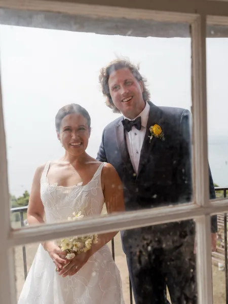 Couple portrait of bride and groom smiling through a window, bride holding bouquet, with ocean and balcony railing behind them