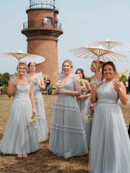 Bridesmaid group photo in light blue bridesmaid dresses, holding white paper parasols and small bouquets by a brick lighthouse tower on grass
