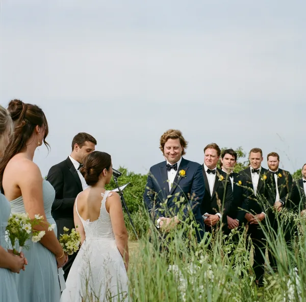Ceremony moment at an outdoor wedding ceremony with bride and groom, wedding party in tuxedos and blue dresses amid tall grass and wildflowers