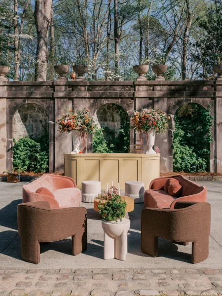 Wedding lounge setup for a cocktail hour lounge arranged against a stone wall with arches on a cobblestone patio, accented with florals.