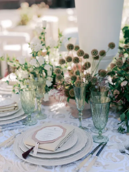 Wedding place setting on lace table linens with pressed glass goblets, set amid garden greenery and white chairs in daylight.