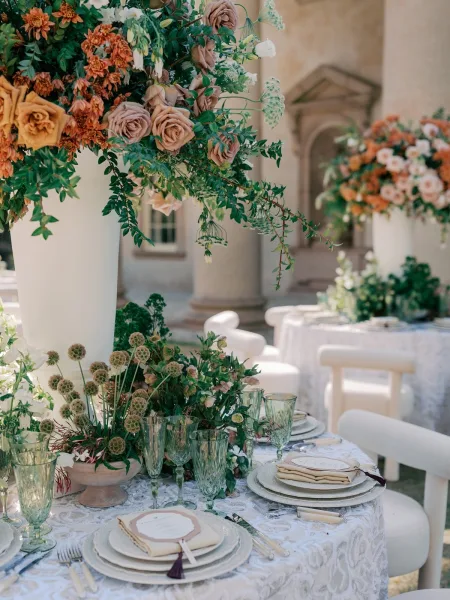 Reception tablescape for an outdoor wedding table with tall floral centerpieces, set in a courtyard with a stone wall and garden greenery.