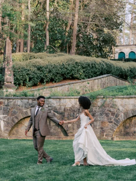 Couple portrait of the bride and groom walking hand in hand by a stone wall and garden arches, highlighted by the groom's boutonniere.