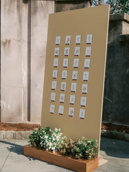 Seating chart display serving as an escort card display on a wooden base, set against a stone wall with outdoor trees and concrete pavement.