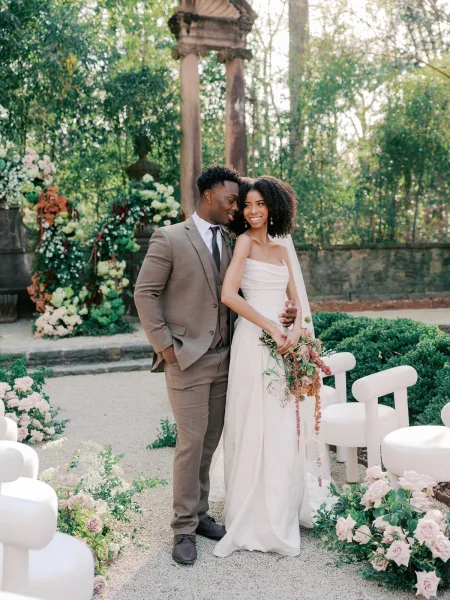 Couple portrait of a bride and groom standing together, she holds a cascading rose and greenery bouquet amid stone columns and garden trees.