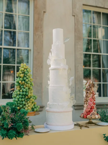 Wedding cake, a white fondant cake, is displayed on a gold cake stand against a stone wall and large windows.