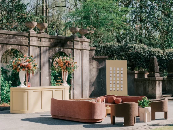 Wedding lounge setup for a cocktail hour lounge featuring floral arrangements in a garden setting against a stone wall with ivy-clad arches.