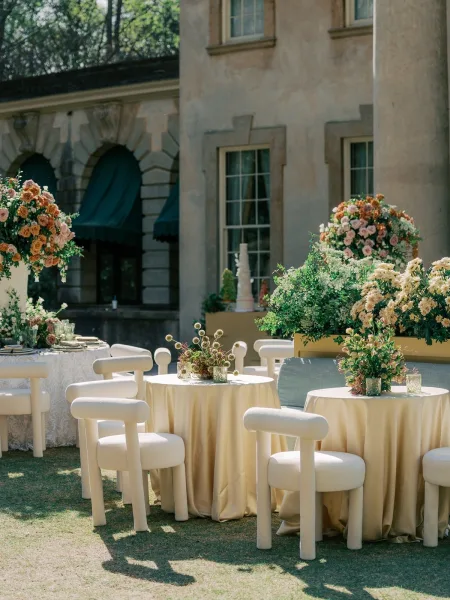 cocktail hour seating for outdoor reception seating, round tables glow with votive candles on a lawn before a stone building with green awnings