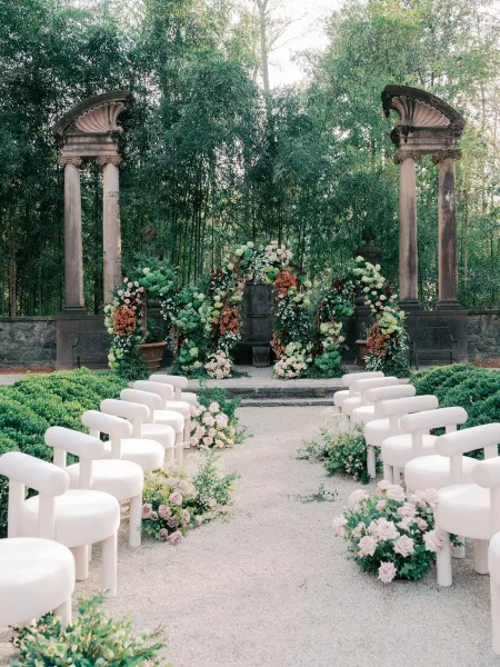 Ceremony setup showcasing refined outdoor ceremony decor, with roses framing the aisle before bamboo trees and a stone wall.