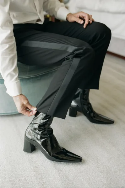 Groom getting ready, pulling on black patent leather boots with tuxedo pants satin stripe beside teal velvet ottoman in hotel room