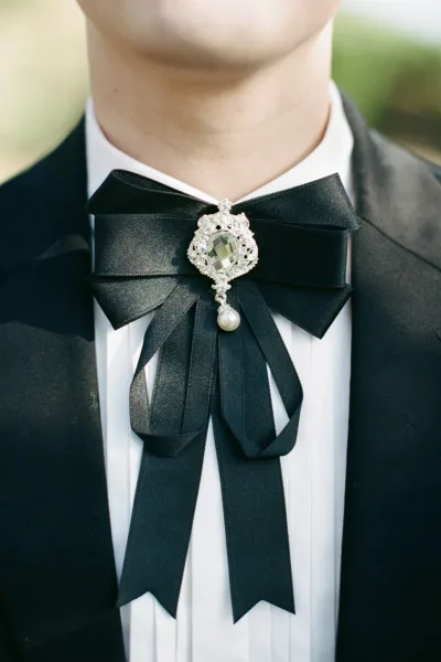 Groom bow tie in black satin with a crystal brooch and pearl drop on a white dress shirt and black tuxedo jacket, greenery blurred behind
