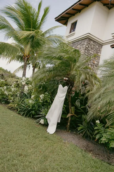 Wedding dress, strapless wedding dress in satin with a ruched bodice on a hanger outside by palm trees and a stucco wall under cloudy sky