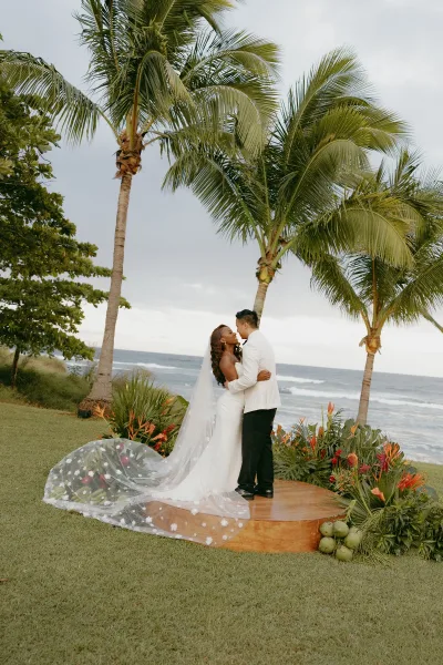 Wedding kiss portrait of bride and groom kissing with a long veil train on a wooden platform, palm trees and ocean behind them