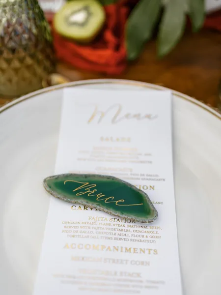Wedding place card on a green agate place card with gold calligraphy, set on a white plate with red napkin, kiwi and pineapple on wood table