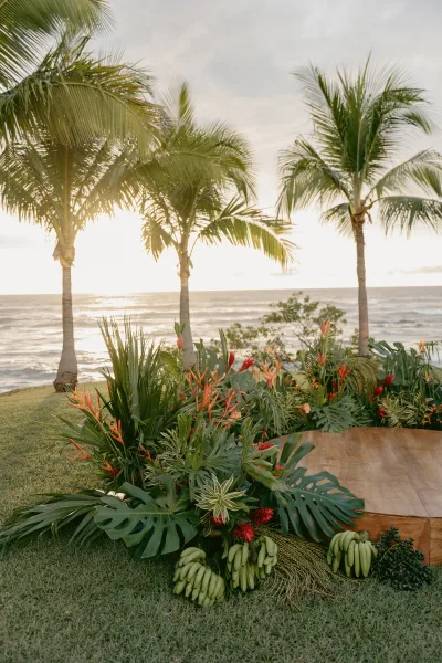 Ceremony backdrop with tropical wedding ceremony florals and palm leaves on a wooden platform, set by oceanfront palms at sunset