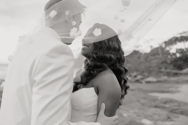 Couple portrait in a black and white wedding portrait style, bride and groom under veil smiling close together on a hillside with trees and sky