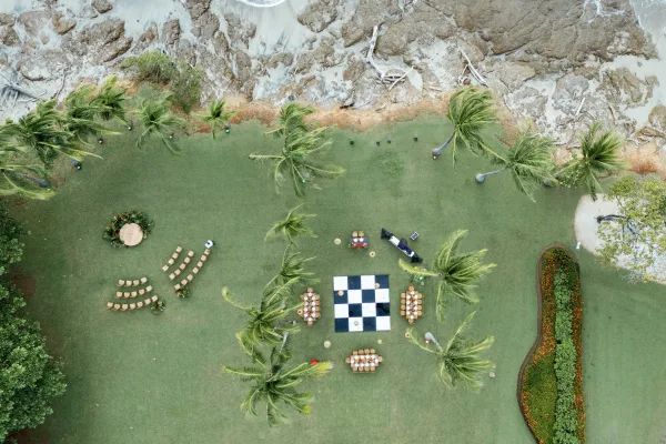 Outdoor wedding reception with palm trees, round sweetheart table, and black-and-white dance floor beside a coastal lawn and ocean edge