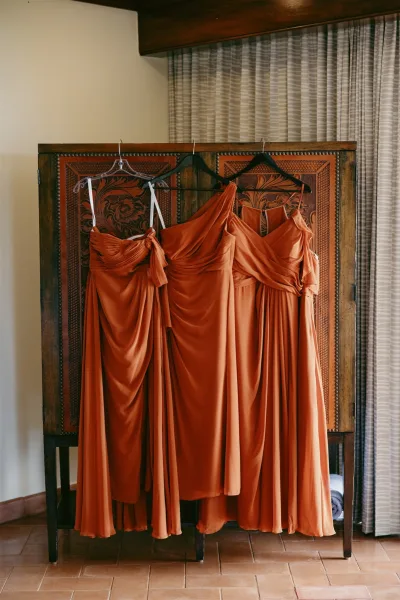 Bridesmaid dresses in rust tones hang on dress hangers, satin fabric draping neatly against a wood cabinet with curtains behind