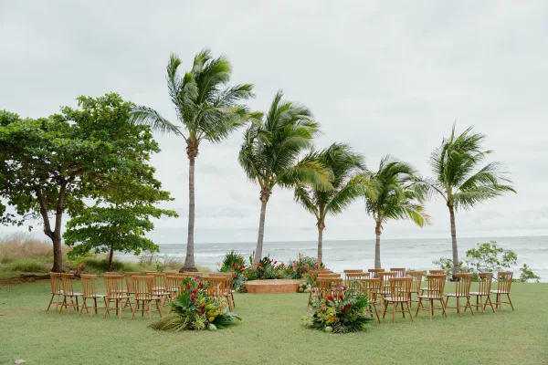 Beach ceremony setup with oceanfront wedding ceremony seating around a wood platform, framed by tropical florals and palms under a cloudy sky