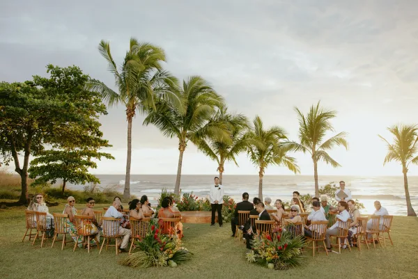 Outdoor wedding ceremony with wood ceremony chairs in a semicircle, floral aisle arrangements, and a groom in white by the ocean and palm trees