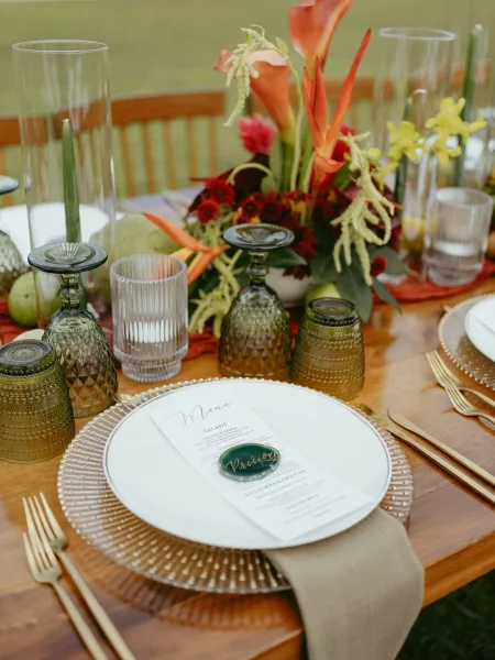 Reception tablescape with wedding table centerpiece of orange calla lilies, red blooms, and taper candles in glass hurricanes on a wood table